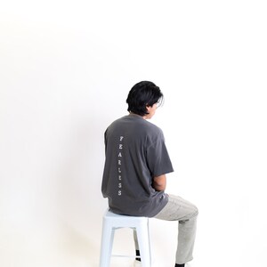May include: A person wearing a charcoal grey t-shirt with the word "FEARLESS" printed vertically down the back. They are sitting on a white metal stool and wearing light grey pants and white sneakers.
