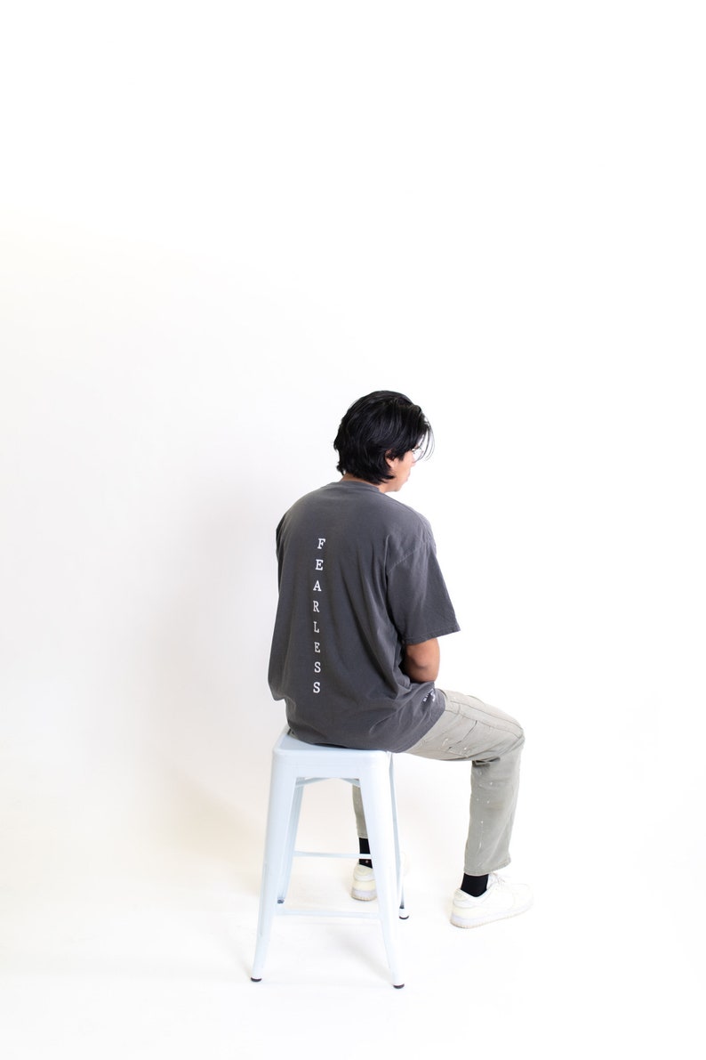 May include: A person wearing a charcoal grey t-shirt with the word "FEARLESS" printed vertically down the back. They are sitting on a white metal stool and wearing light grey pants and white sneakers.