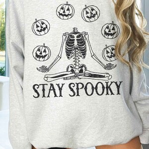 May include: A gray sweatshirt with a skeleton meditating and holding pumpkins. The text "STAY SPOOKY" is printed below the skeleton.