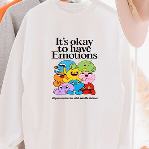 Peut inclure: Sweat-shirt blanc avec un motif de personnages de dessins anim&eacute;s color&eacute;s et le texte "It's okay to have Emotions" et "all your emotions are valid... even the sad ones".