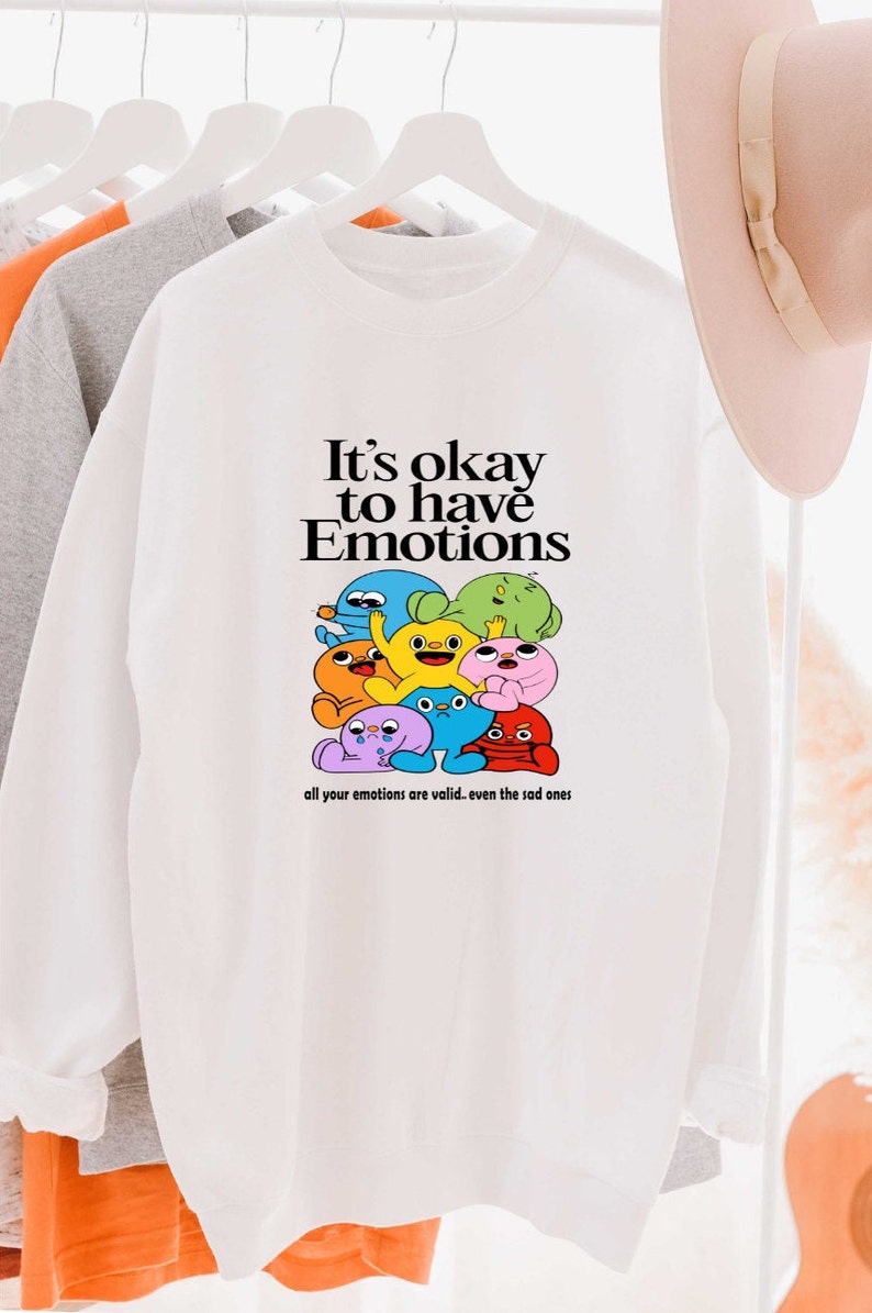 Peut inclure: Sweat-shirt blanc avec un motif de personnages de dessins anim&eacute;s color&eacute;s et le texte "It's okay to have Emotions" et "all your emotions are valid... even the sad ones".