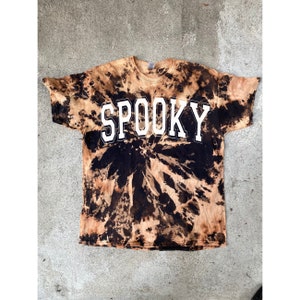以下が含まれることがあります： 黒と茶色のタイダイ染めのTシャツで、前面に白い文字で「SPOOKY」とプリントされています。