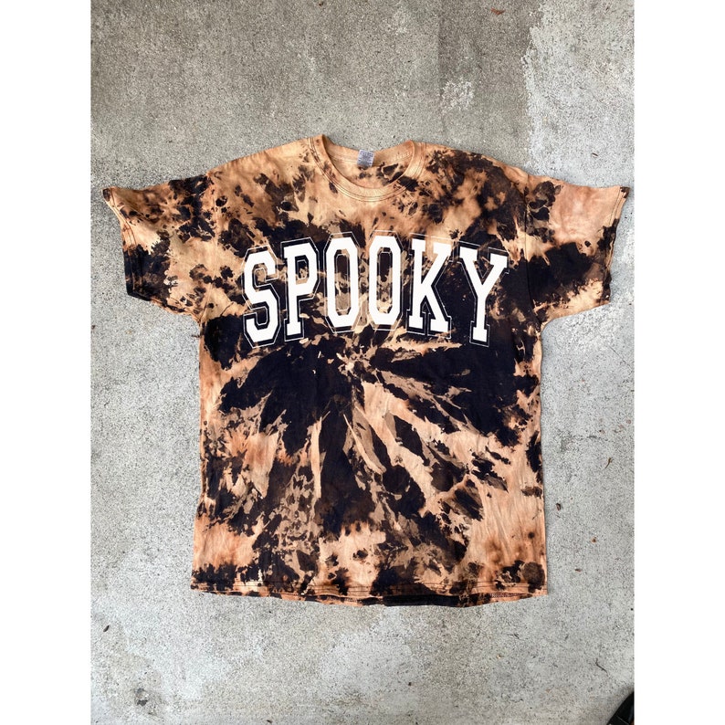 以下が含まれることがあります： 黒と茶色のタイダイ染めのTシャツで、前面に白い文字で「SPOOKY」とプリントされています。