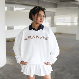Puede incluir: Sudadera blanca con el texto "JESUS IS KING" impreso en marr&oacute;n en la parte delantera. La sudadera la lleva puesta una persona con el pelo oscuro y se combina con una falda blanca.