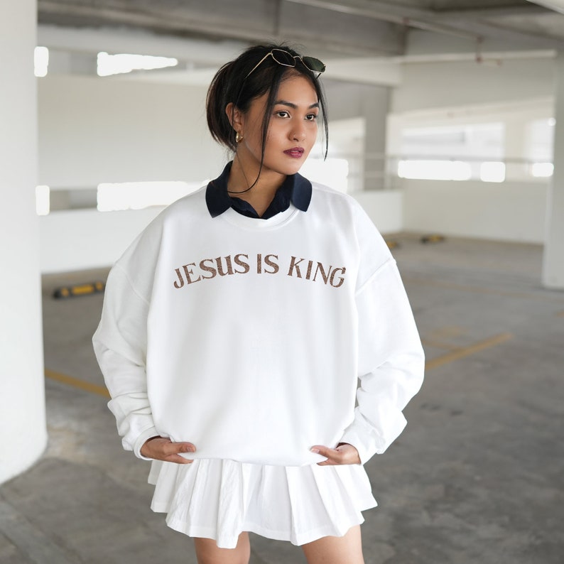 Puede incluir: Sudadera blanca con el texto "JESUS IS KING" impreso en marr&oacute;n en la parte delantera. La sudadera la lleva puesta una persona con el pelo oscuro y se combina con una falda blanca.