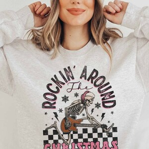 K&ouml;nnte beinhalten: Ein graues Sweatshirt mit einem Aufdruck eines Skeletts, das Gitarre spielt, mit dem Text "Rockin' Around the Christmas Tree" in Pink und Schwarz.