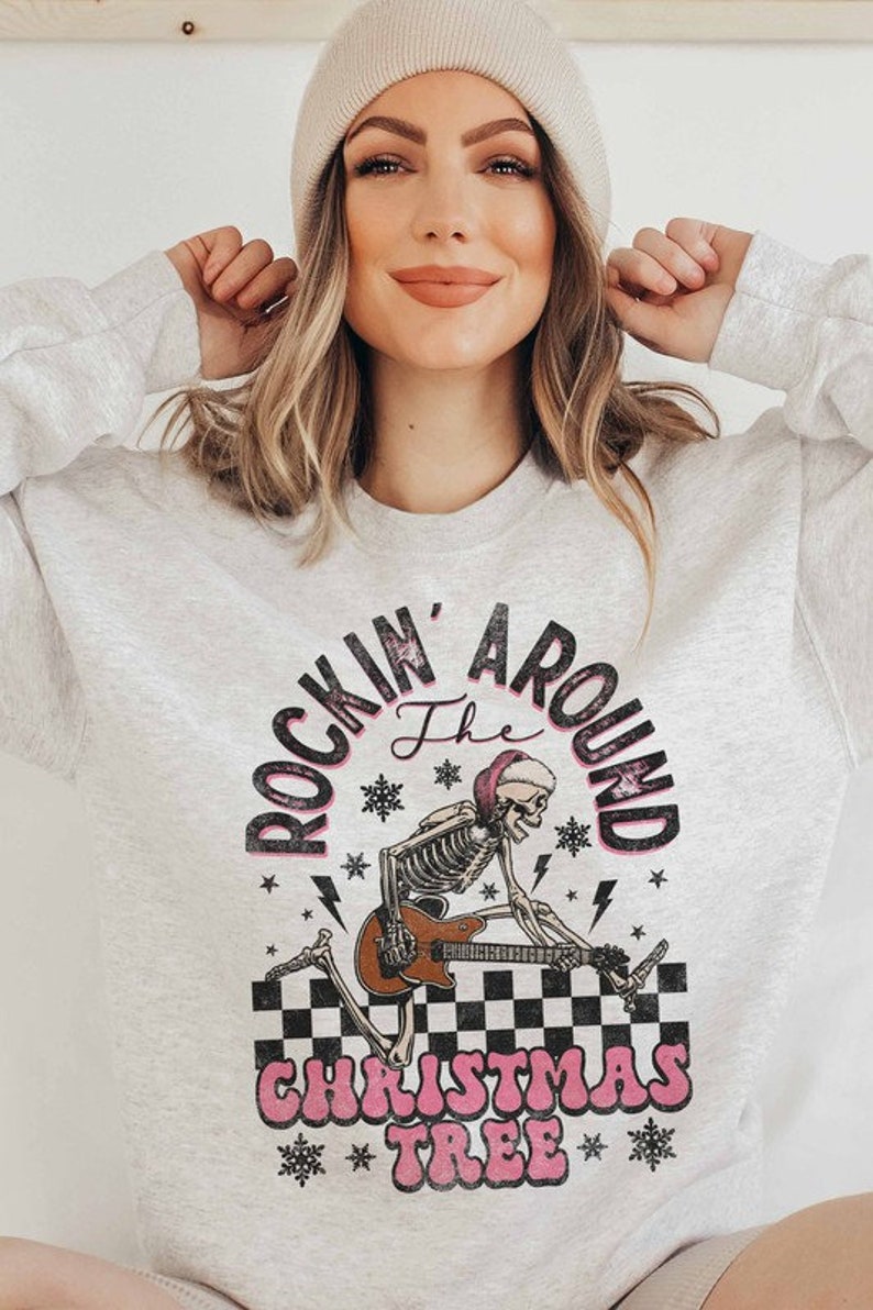 K&ouml;nnte beinhalten: Ein graues Sweatshirt mit einem Aufdruck eines Skeletts, das Gitarre spielt, mit dem Text "Rockin' Around the Christmas Tree" in Pink und Schwarz.
