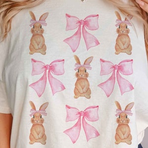 Pu&ograve; includere: Una maglietta bianca con un motivo ripetitivo di fiocchi rosa e coniglietti marroni che indossano fiocchi rosa.
