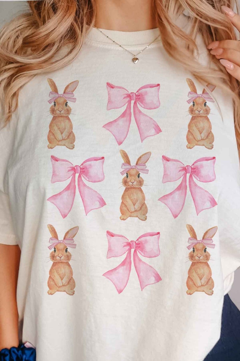 Pu&ograve; includere: Una maglietta bianca con un motivo ripetitivo di fiocchi rosa e coniglietti marroni che indossano fiocchi rosa.