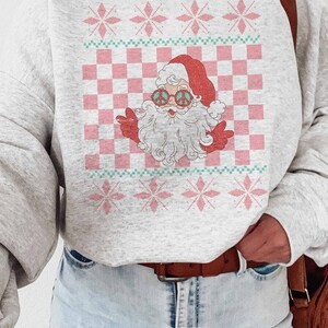 K&ouml;nnte beinhalten: Ein grauer Pullover mit einem rosa und gr&uuml;nen Karomuster und einem Cartoon-Weihnachtsmann, der eine Peace-Sign-Sonnenbrille tr&auml;gt. Der Weihnachtsmann tr&auml;gt einen roten Hut und einen wei&szlig;en Bart.