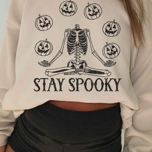 May include: A beige sweatshirt with a skeleton meditating with pumpkins around it. The text "STAY SPOOKY" is printed below the skeleton.