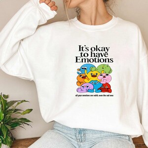 Peut inclure: Sweat-shirt blanc avec un motif de personnages de dessins anim&eacute;s color&eacute;s et le texte "It's okay to have Emotions. All your emotions are valid. Even the sad ones."