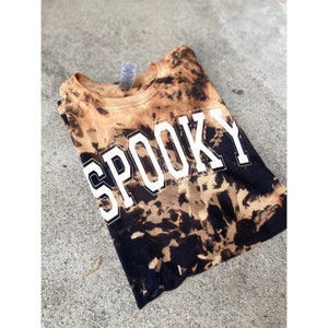 以下が含まれることがあります： 黒と茶色のタイダイ染めのTシャツで、前面に白い文字で「SPOOKY」とプリントされています。