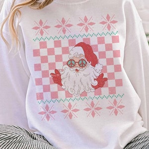 K&ouml;nnte beinhalten: Wei&szlig;er Weihnachtspullover mit einem rosa-gr&uuml;nen Karomuster und einem Cartoon-Weihnachtsmann mit Peace-Zeichen-Sonnenbrille. Der Pullover hat ein rosa Schneeflockenmuster.