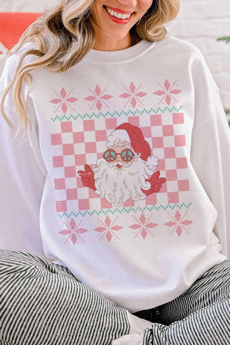 K&ouml;nnte beinhalten: Wei&szlig;er Weihnachtspullover mit einem rosa-gr&uuml;nen Karomuster und einem Cartoon-Weihnachtsmann mit Peace-Zeichen-Sonnenbrille. Der Pullover hat ein rosa Schneeflockenmuster.