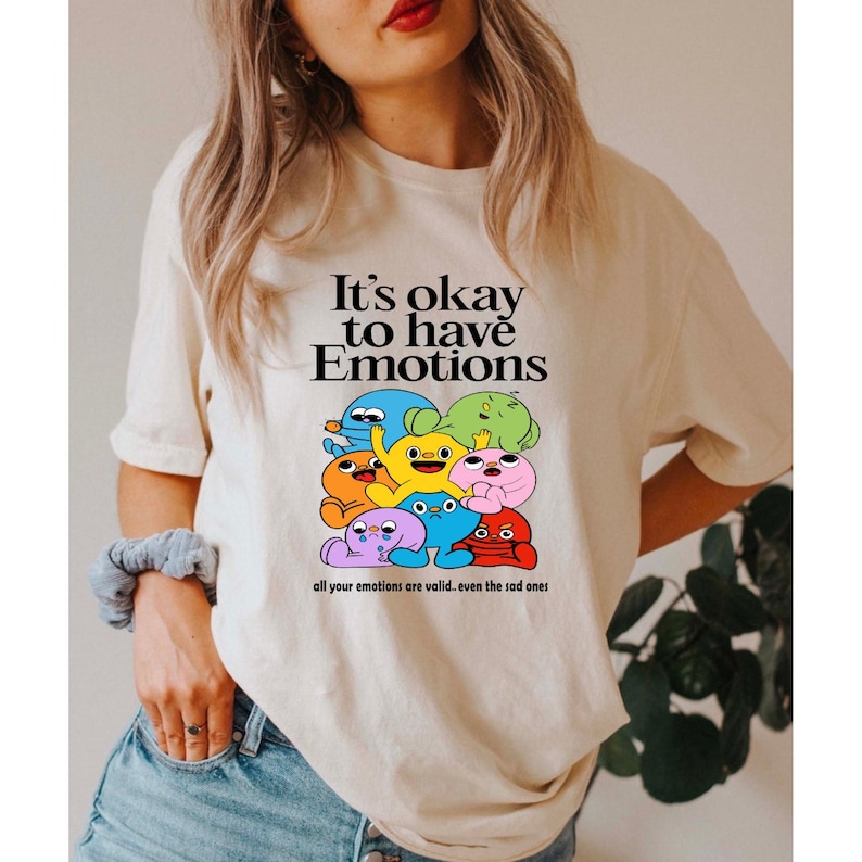 Op de afbeelding: Een wit T-shirt met een grafische print van kleurrijke cartoonfiguren. De tekst "It's okay to have Emotions" is boven de figuren gedrukt. De tekst "all your emotions are valid...even the sad ones" is onder de figuren gedrukt.