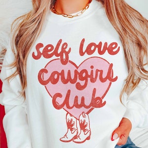 K&ouml;nnte beinhalten: Wei&szlig;er Pullover mit einem rosa Herz und rotem Text, der "self love cowgirl club" lautet. Das Herz hat unten ein Paar rote Cowboystiefel.