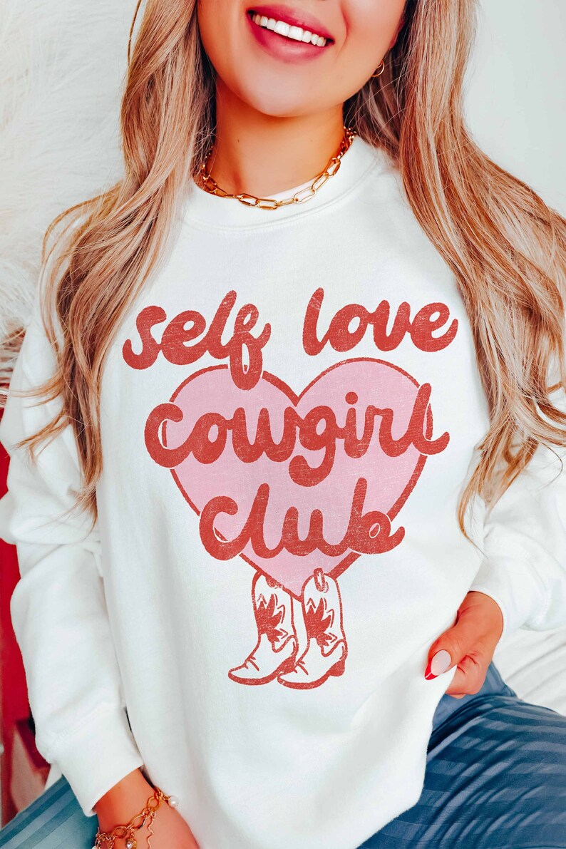 K&ouml;nnte beinhalten: Wei&szlig;er Pullover mit einem rosa Herz und rotem Text, der "self love cowgirl club" lautet. Das Herz hat unten ein Paar rote Cowboystiefel.