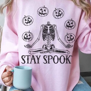 May include: A pink sweatshirt with a skeleton meditating with pumpkins around it. The text "STAY SPOOK" is printed below the skeleton.