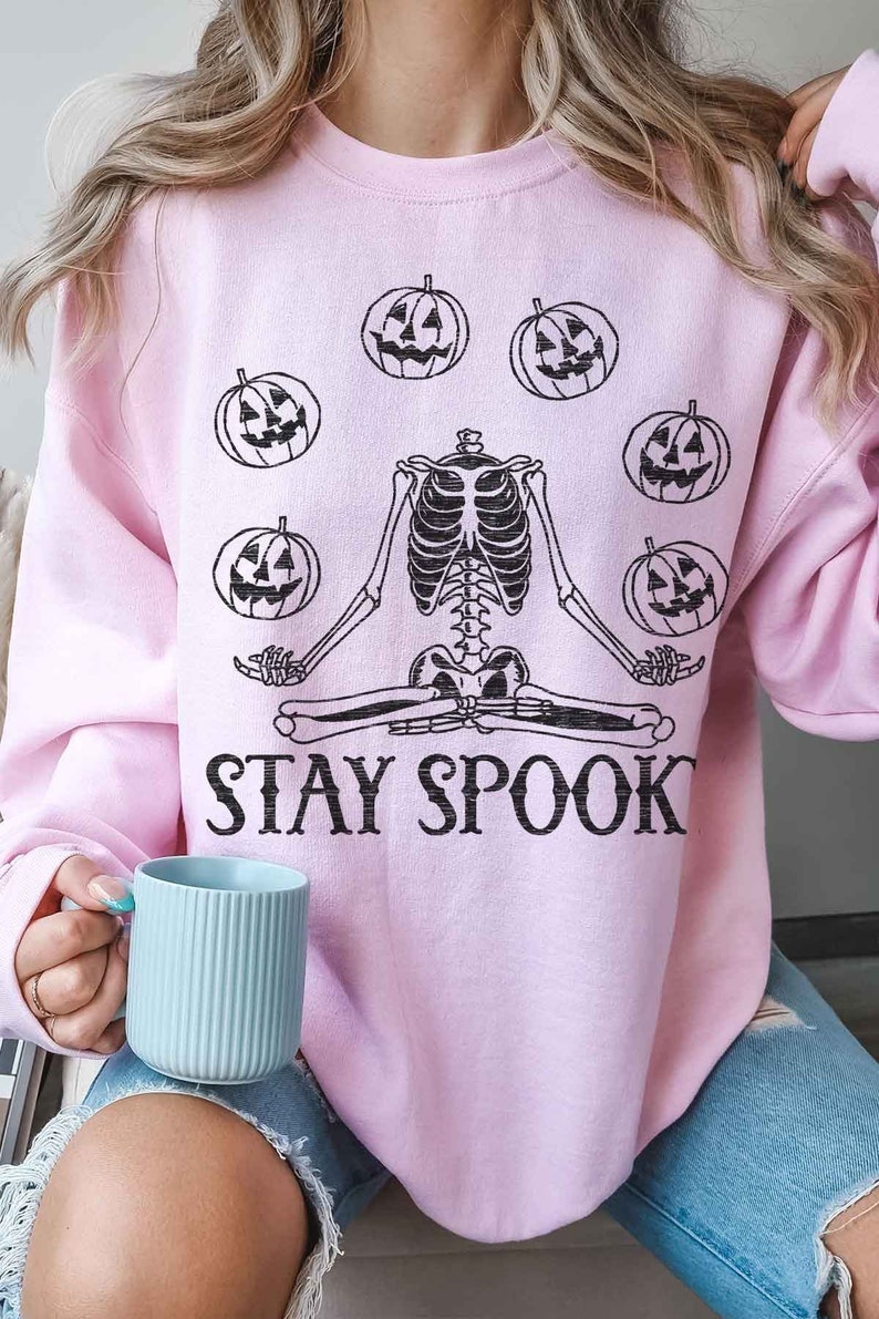 May include: A pink sweatshirt with a skeleton meditating with pumpkins around it. The text "STAY SPOOK" is printed below the skeleton.