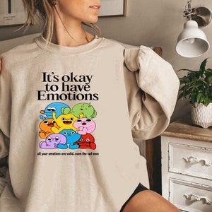 Peut inclure: Un sweat-shirt beige avec un imprim&eacute; graphique de personnages de dessins anim&eacute;s color&eacute;s. Le texte sur le sweat-shirt est "It's okay to have Emotions all your emotions are valid.. even the sad ones."