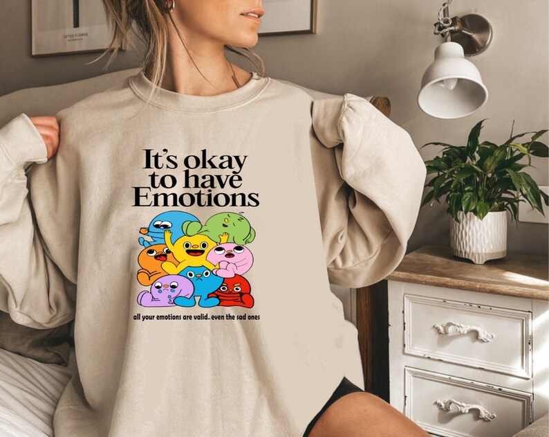 Peut inclure: Un sweat-shirt beige avec un imprim&eacute; graphique de personnages de dessins anim&eacute;s color&eacute;s. Le texte sur le sweat-shirt est "It's okay to have Emotions all your emotions are valid.. even the sad ones."
