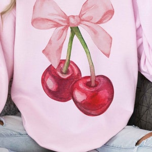 May include: A pink sweatshirt with a watercolor illustration of two cherries with a pink bow on top.