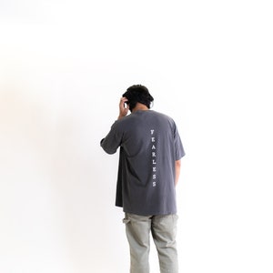 May include: A person wearing a dark gray t-shirt with the word "FEARLESS" printed vertically down the back. The person is wearing light green cargo pants and white sneakers.