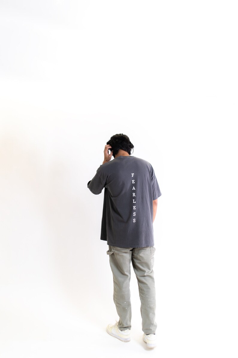 May include: A person wearing a dark gray t-shirt with the word "FEARLESS" printed vertically down the back. The person is wearing light green cargo pants and white sneakers.