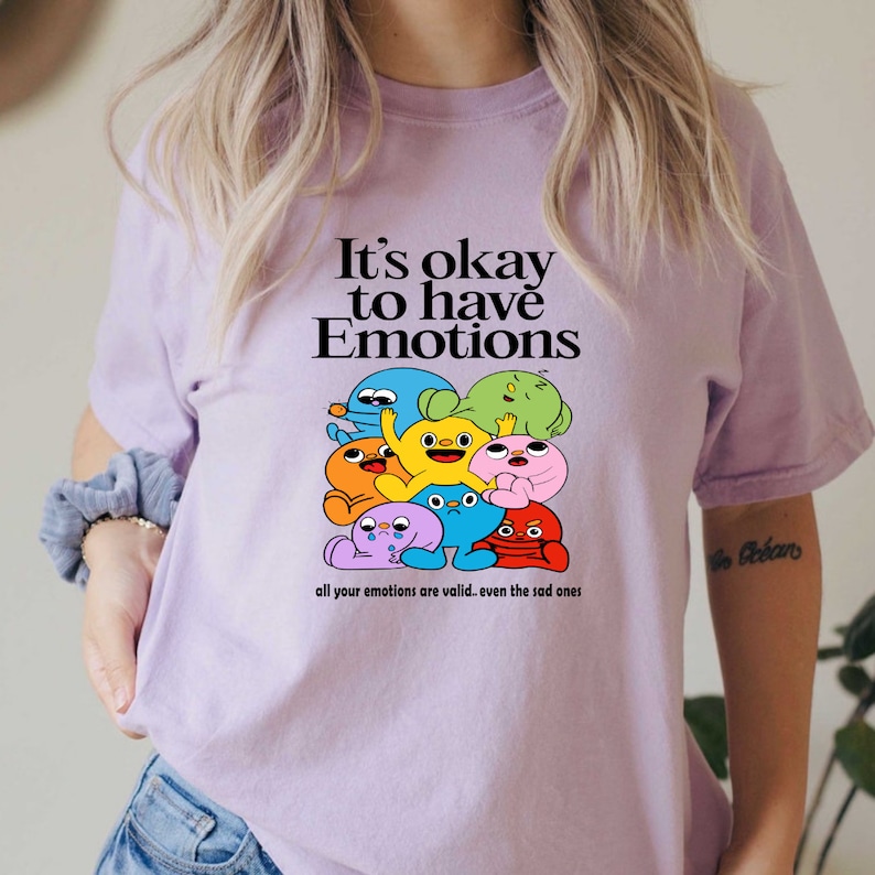 Op de afbeelding: Een lavendelkleurig T-shirt met een afbeelding van kleurrijke cartoonfiguren en de tekst "It's okay to have Emotions. All your emotions are valid... even the sad ones."