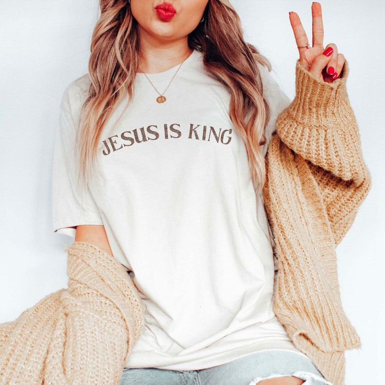 Puede incluir: Una camiseta blanca con el texto "JESUS IS KING" impreso en negro. La camiseta la lleva puesta una persona con el pelo largo y rubio y un c&aacute;rdigan marr&oacute;n.