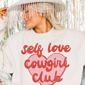 K&ouml;nnte beinhalten: Ein wei&szlig;er Pullover mit einem rosa Herz und rotem Text, der "self love cowgirl club" lautet. Das Herz hat zwei rote Cowboystiefel unten.