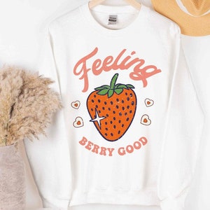 May include: White long-sleeved sweatshirt with a pink and orange graphic of a strawberry with the text "Feeling Berry Good".