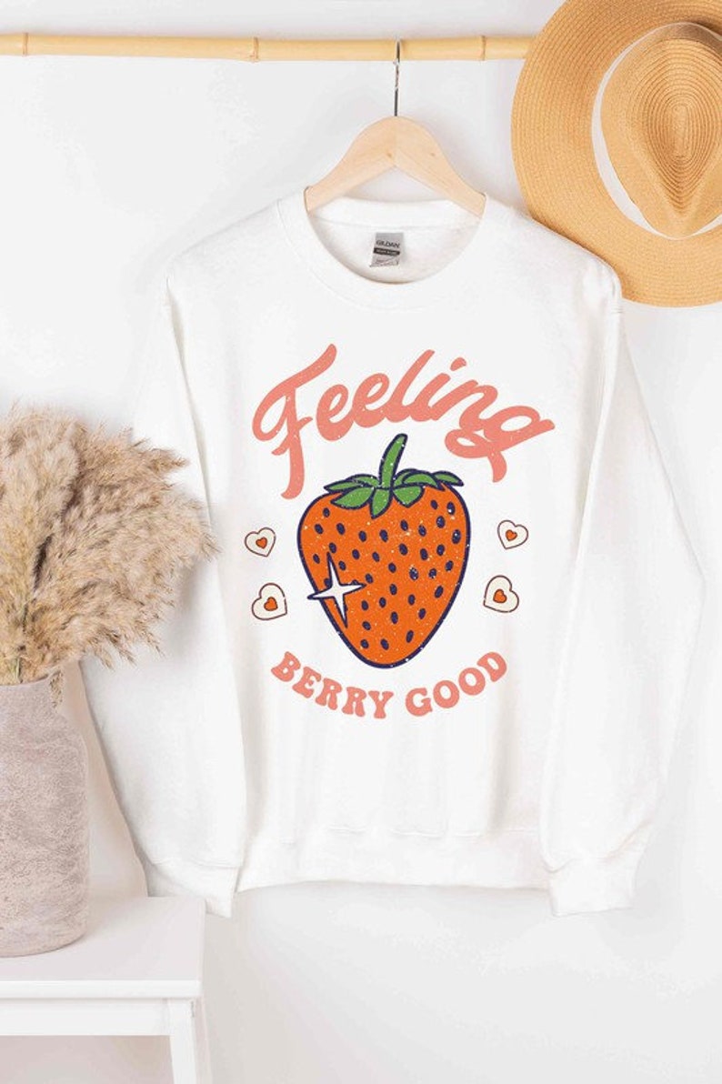 May include: White long-sleeved sweatshirt with a pink and orange graphic of a strawberry with the text "Feeling Berry Good".