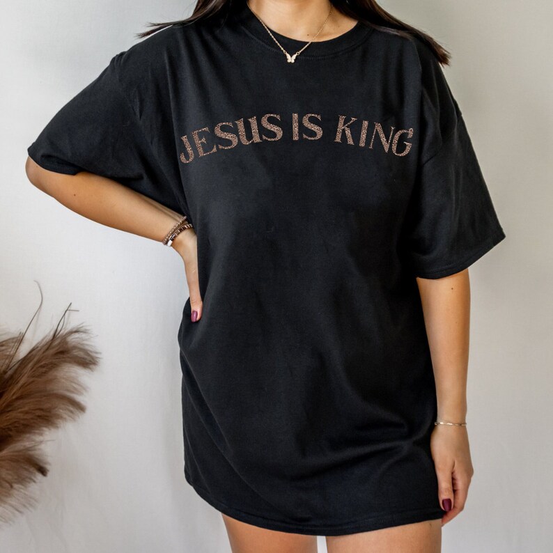Puede incluir: Camiseta negra de gran tama&ntilde;o con el texto "JESUS IS KING" en letras de purpurina dorada.