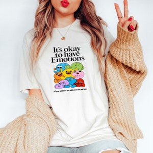 Op de afbeelding: Wit T-shirt met een kleurrijke afbeelding van cartoonfiguren en de tekst "It's okay to have Emotions all your emotions are valid. even the sad ones."