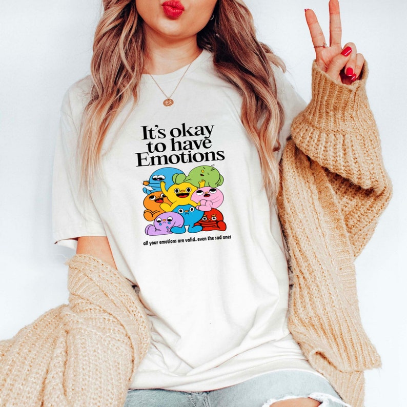 Op de afbeelding: Wit T-shirt met een kleurrijke afbeelding van cartoonfiguren en de tekst "It's okay to have Emotions all your emotions are valid. even the sad ones."