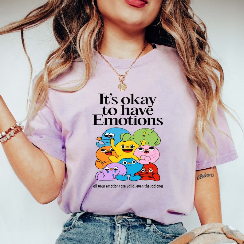 Op de afbeelding: Een lichtpaarse t-shirt met een afbeelding van kleurrijke cartoonfiguren en de tekst "It's okay to have Emotions. All your emotions are valid. Even the sad ones."