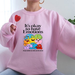Peut inclure: Sweat-shirt rose avec un motif de personnages de dessins anim&eacute;s color&eacute;s et le texte "It's okay to have Emotions all your emotions are valid, even the sad ones."