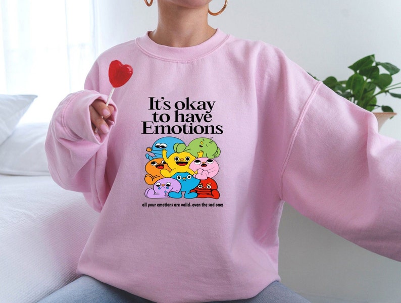 Peut inclure: Sweat-shirt rose avec un motif de personnages de dessins anim&eacute;s color&eacute;s et le texte "It's okay to have Emotions all your emotions are valid, even the sad ones."