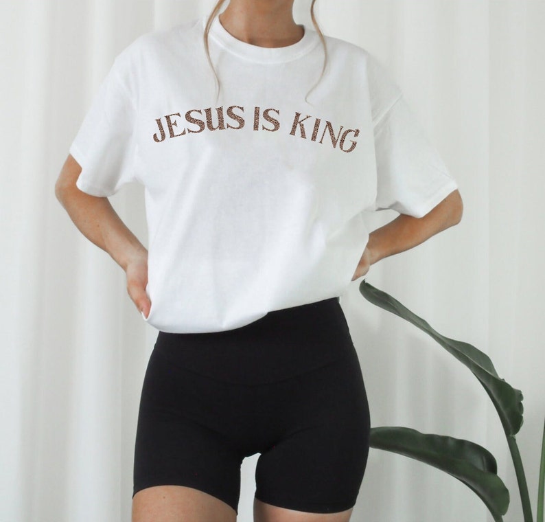 Puede incluir: Camiseta blanca con el texto "JESUS IS KING" en letras marrones. La camiseta la lleva puesta una persona que lleva pantalones cortos de ciclista negros.