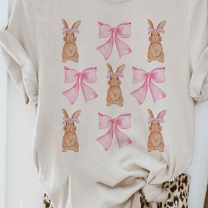 Pu&ograve; includere: Una maglietta beige con un motivo ripetitivo di coniglietti marroni che indossano fiocchi rosa.