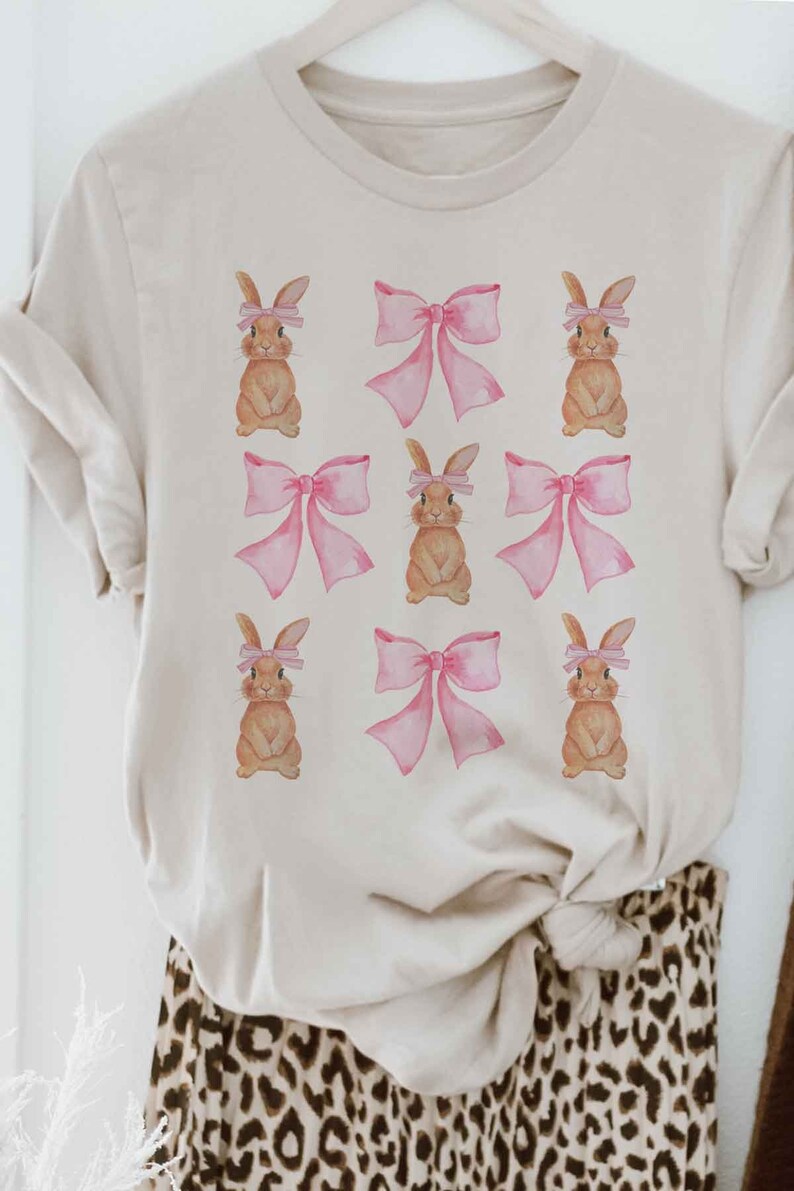 Pu&ograve; includere: Una maglietta beige con un motivo ripetitivo di coniglietti marroni che indossano fiocchi rosa.