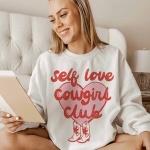 K&ouml;nnte beinhalten: Wei&szlig;er Pullover mit einem rosa Herz-Motiv, auf dem in roten Buchstaben "self love cowgirl club" steht. Zwei rote Cowboystiefel befinden sich am unteren Rand des Herzens.