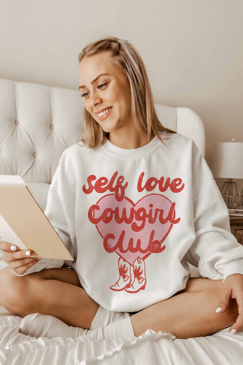 K&ouml;nnte beinhalten: Wei&szlig;er Pullover mit einem rosa Herz-Motiv, auf dem in roten Buchstaben "self love cowgirl club" steht. Zwei rote Cowboystiefel befinden sich am unteren Rand des Herzens.