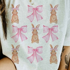 Pu&ograve; includere: Una maglietta bianca con un motivo ripetitivo di conigli marroni che indossano fiocchi rosa.
