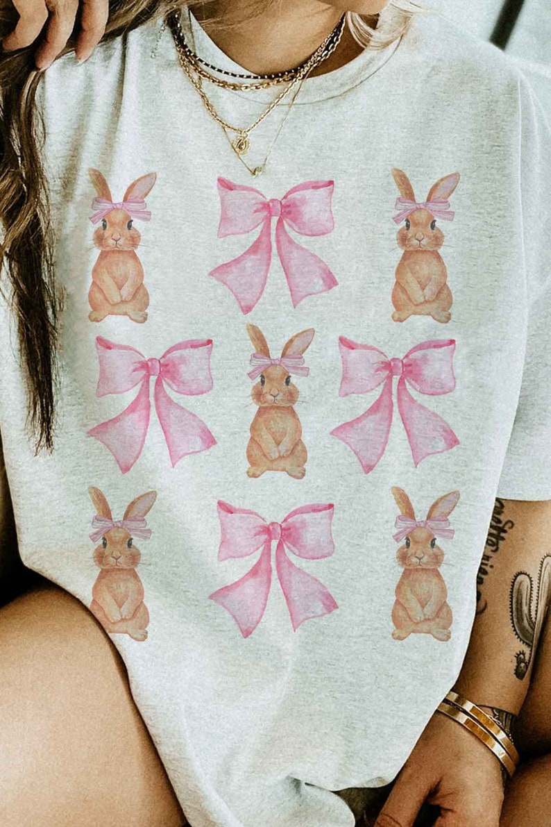 Pu&ograve; includere: Una maglietta bianca con un motivo ripetitivo di conigli marroni che indossano fiocchi rosa.