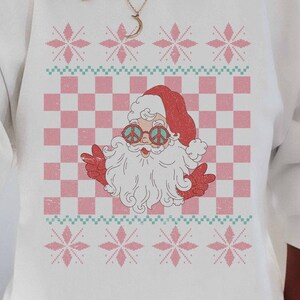 K&ouml;nnte beinhalten: Wei&szlig;er Weihnachtspullover mit einem rosa-gr&uuml;nen Karomuster und einem Cartoon-Weihnachtsmann mit Peace-Zeichen-Sonnenbrille. Der Pullover hat ein festliches Schneeflockenmuster.