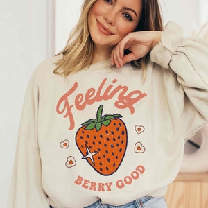 May include: A cream colored sweatshirt with a red strawberry graphic and the text "Feeling Berry Good".