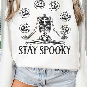 May include: White sweatshirt with a black skeleton meditating with pumpkins around it. The text "STAY SPOOKY" is printed below the skeleton.